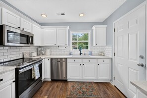 Una cocina con armarios blancos, un microondas de acero inoxidable, un lavavajillas y una ventana con vistas a zonas verdes.