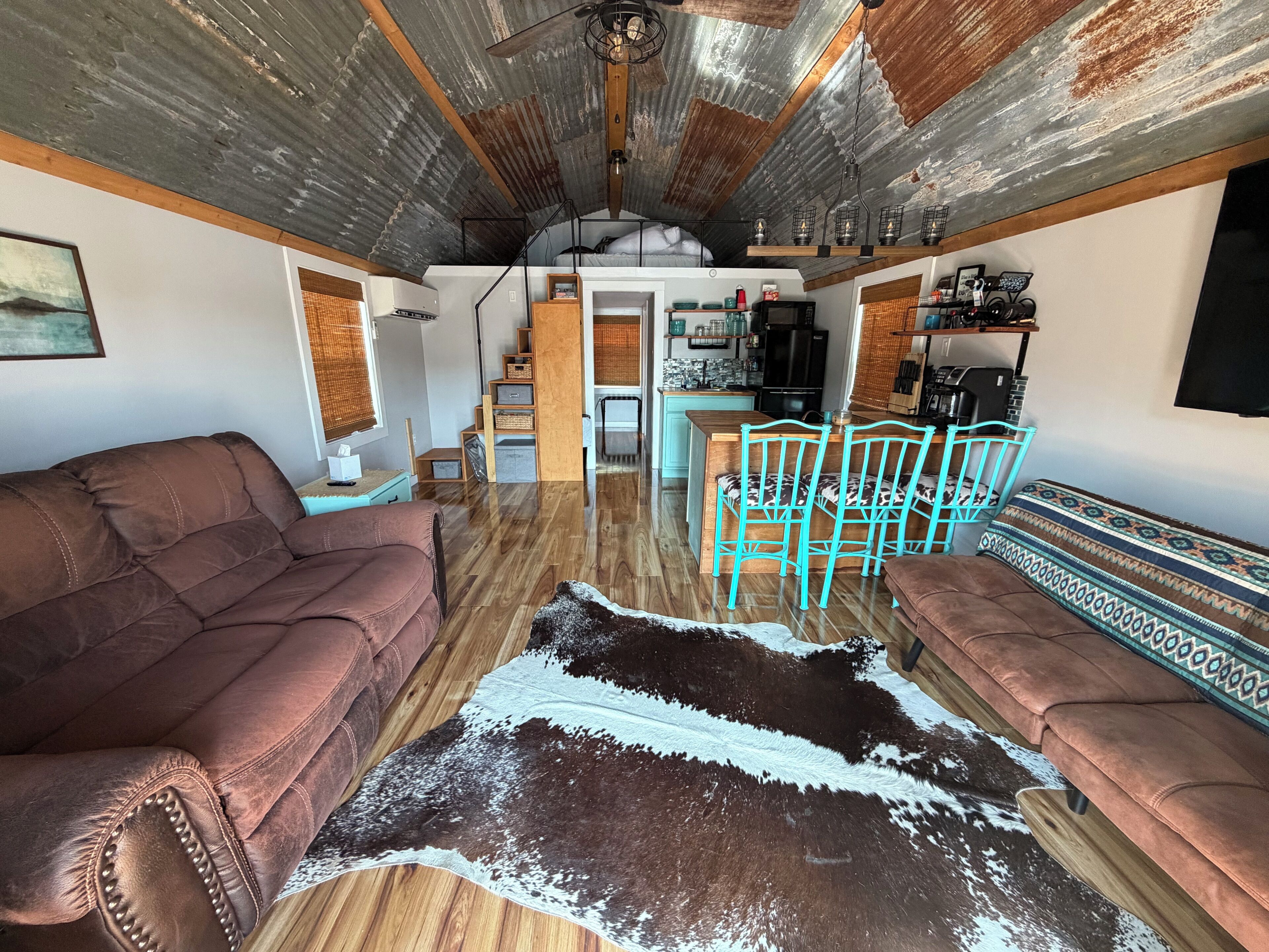 A cozy living area with a brown sofa, teal chairs, a cowhide rug, and a kitchen in the background.