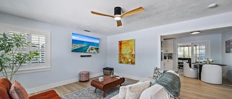 Una sala de estar moderna con un televisor de pantalla plana, un ventilador de techo y una zona de comedor con una mesa y sillas.