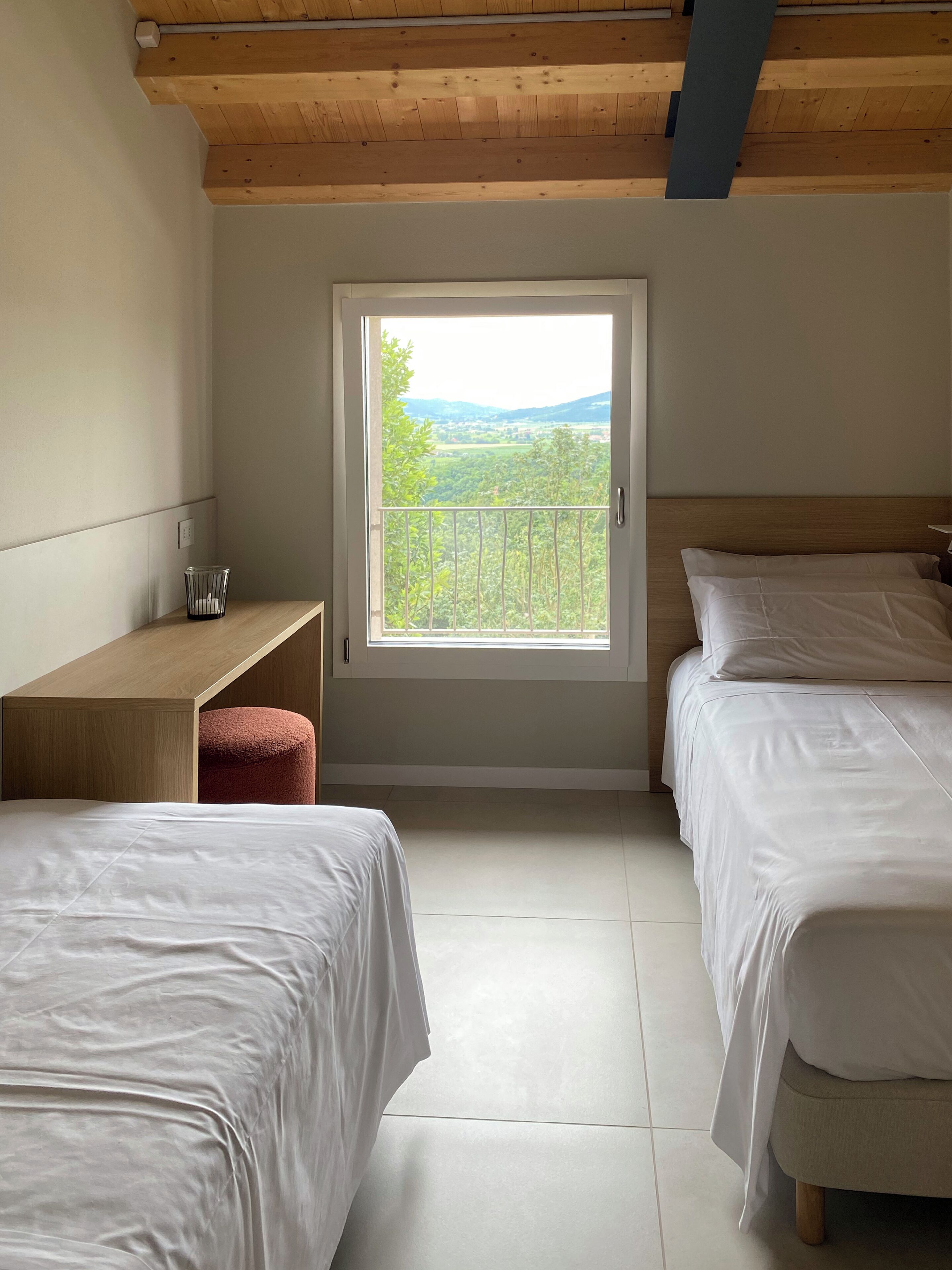 Ein Schlafzimmer mit zwei Betten, einem Holztisch, einem Fenster mit Blick auf Grünflächen und einer geneigten Decke.