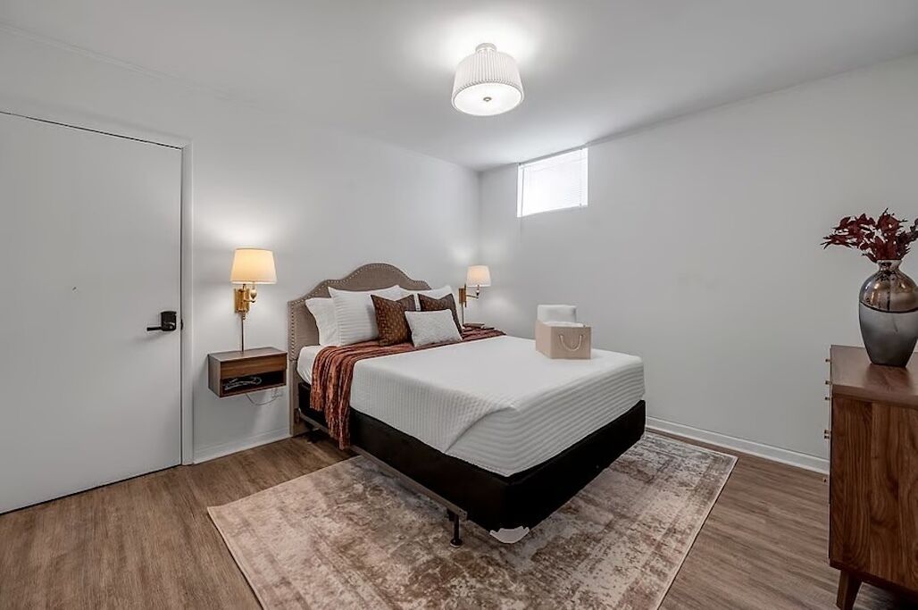 A bedroom with a bed, bedside lamps, a nightstand, a bedside table, a window, and a vase with flowers.