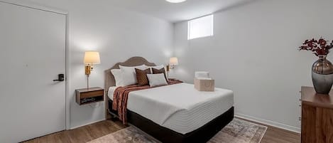 A bedroom with a bed, bedside lamps, a nightstand, a bedside table, a window, and a vase with flowers.