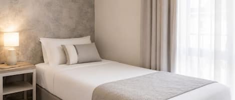 A single bed with a grey blanket, a bedside table with a lamp, and a nightstand with a shelf.