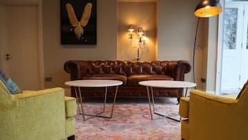 Ein Wohnzimmer mit einem braunen Ledersofa, zwei runden Tischen, einem gelben Sessel, einer Lampe und einem an der Wand befestigten Kunstwerk.