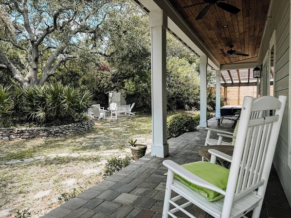 Eine überdachte Veranda mit weißen Stühlen und einem Tisch, eine Steinmauer und ein großer Baum im Hintergrund.