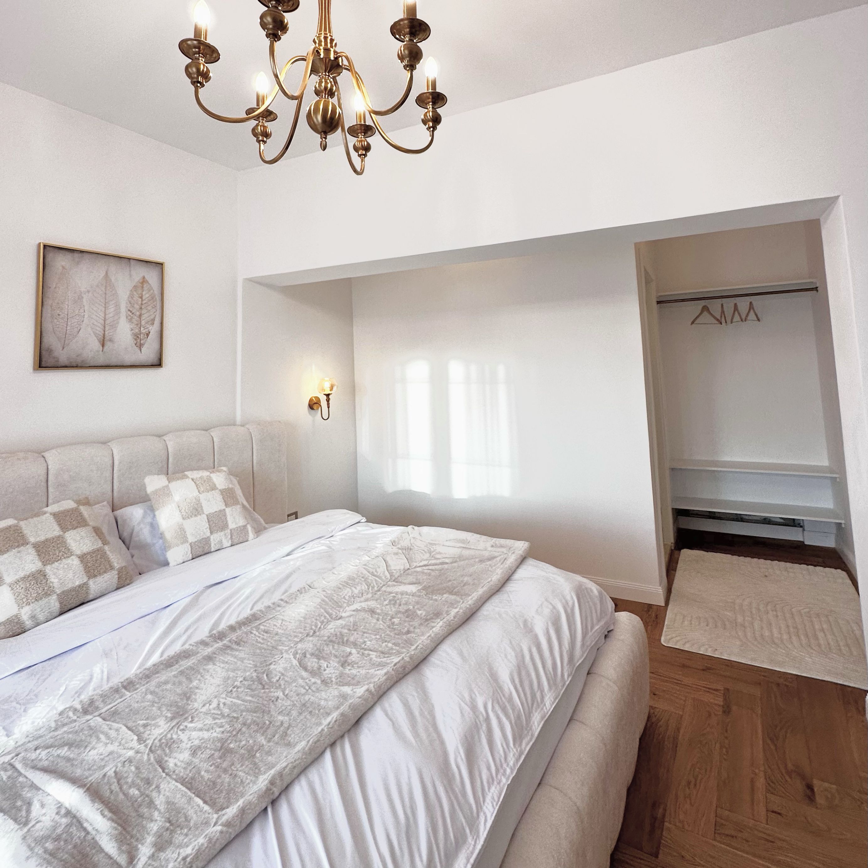Un dormitorio con una cama, una araña, una lámpara de pared, una obra de arte con una hoja enmarcada y un armario con perchas.