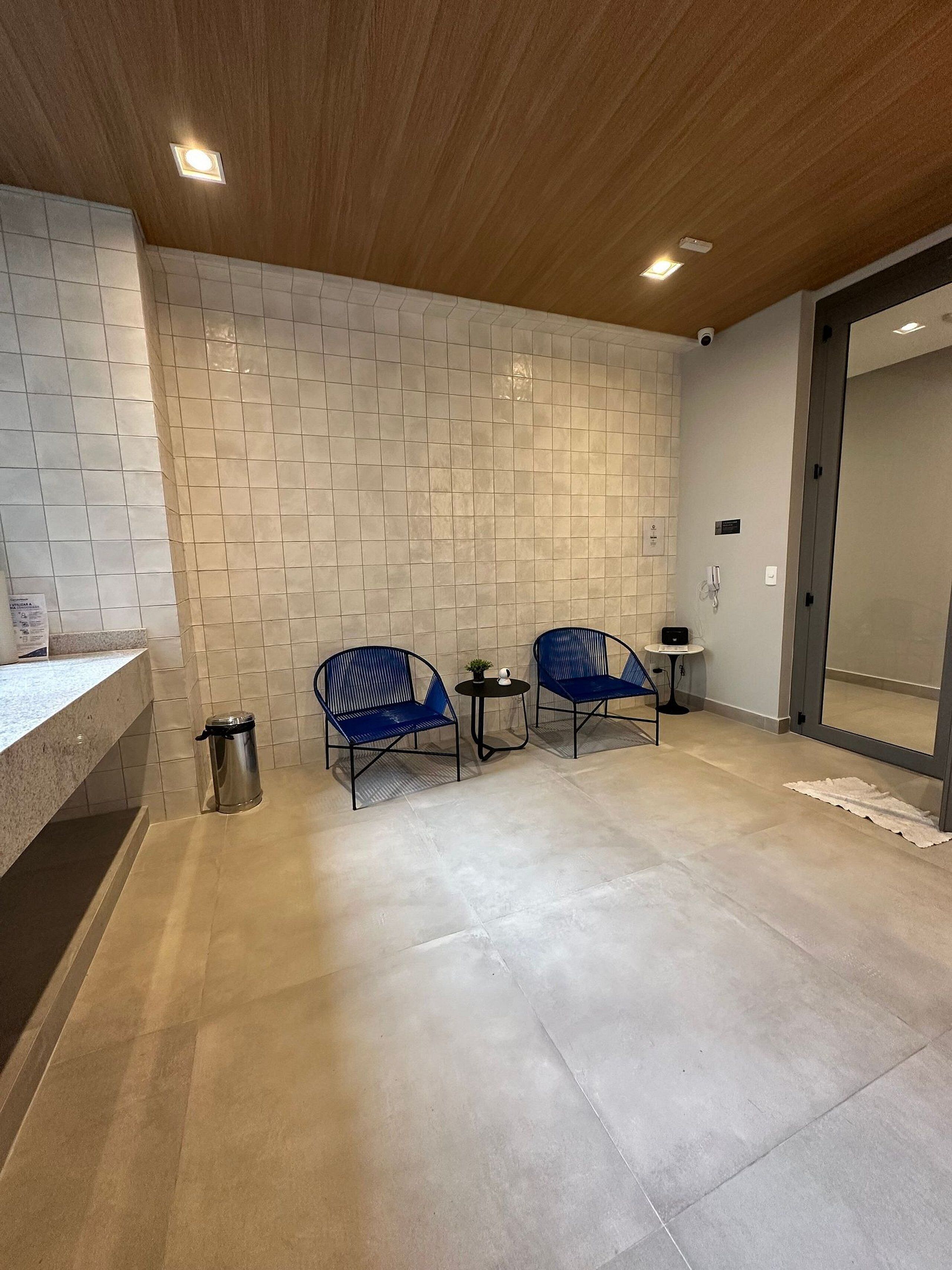 Une salle de bain moderne avec un mur carrelé, un lavabo, deux chaises bleues, une petite table avec une plante et une poubelle.