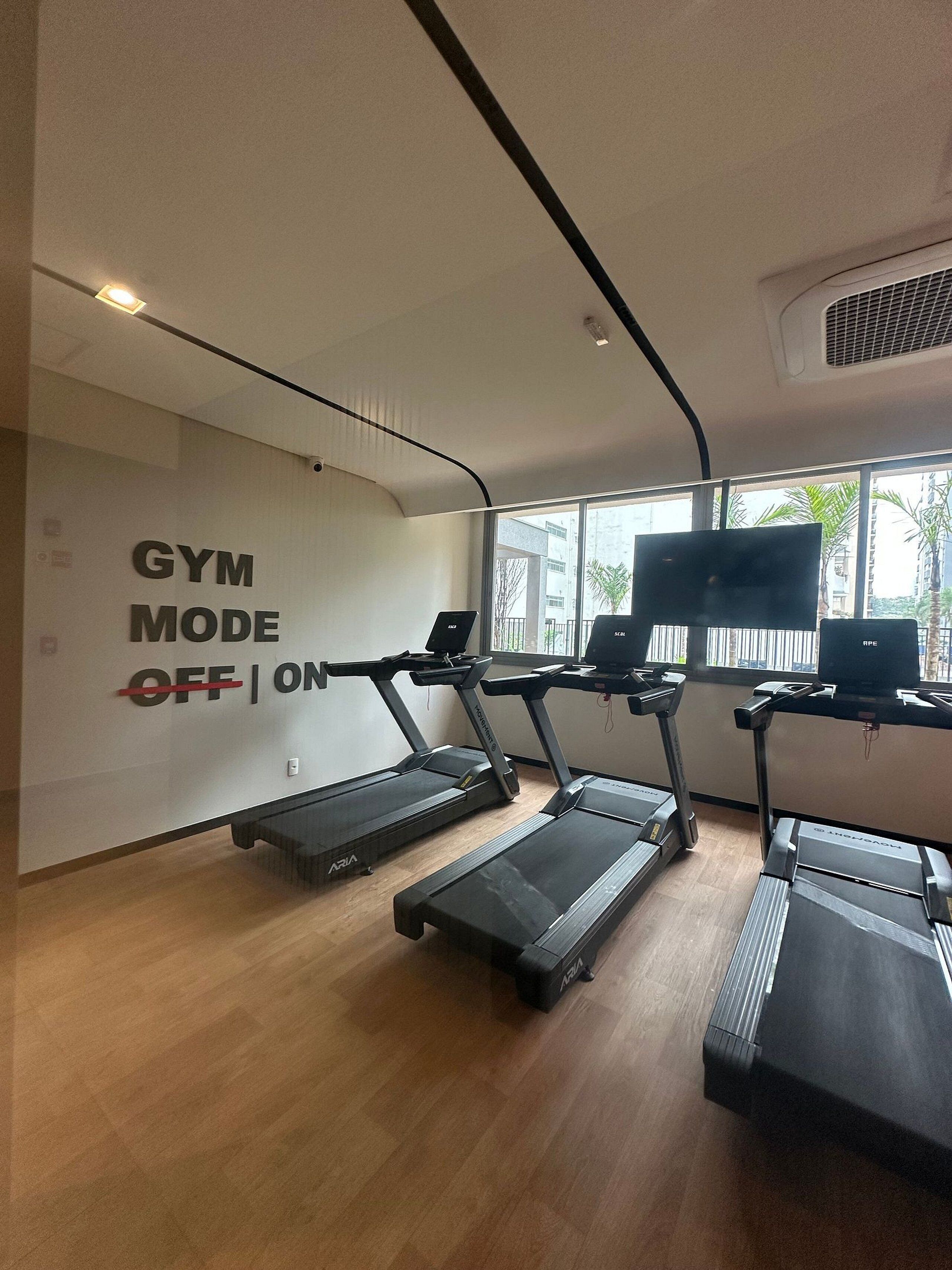 Une salle de sport équipée de tapis de course, un panneau mural indiquant « MODE SALLE DE SPORT : ARRÊTÉ | ACTIVÉ », et de grandes fenêtres offrant une vue sur les bâtiments environnants.