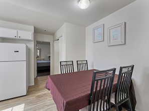 A compact room with a dining table, four chairs, a refrigerator, and a bed in the background. - Jsa & Jsb (Killeen)