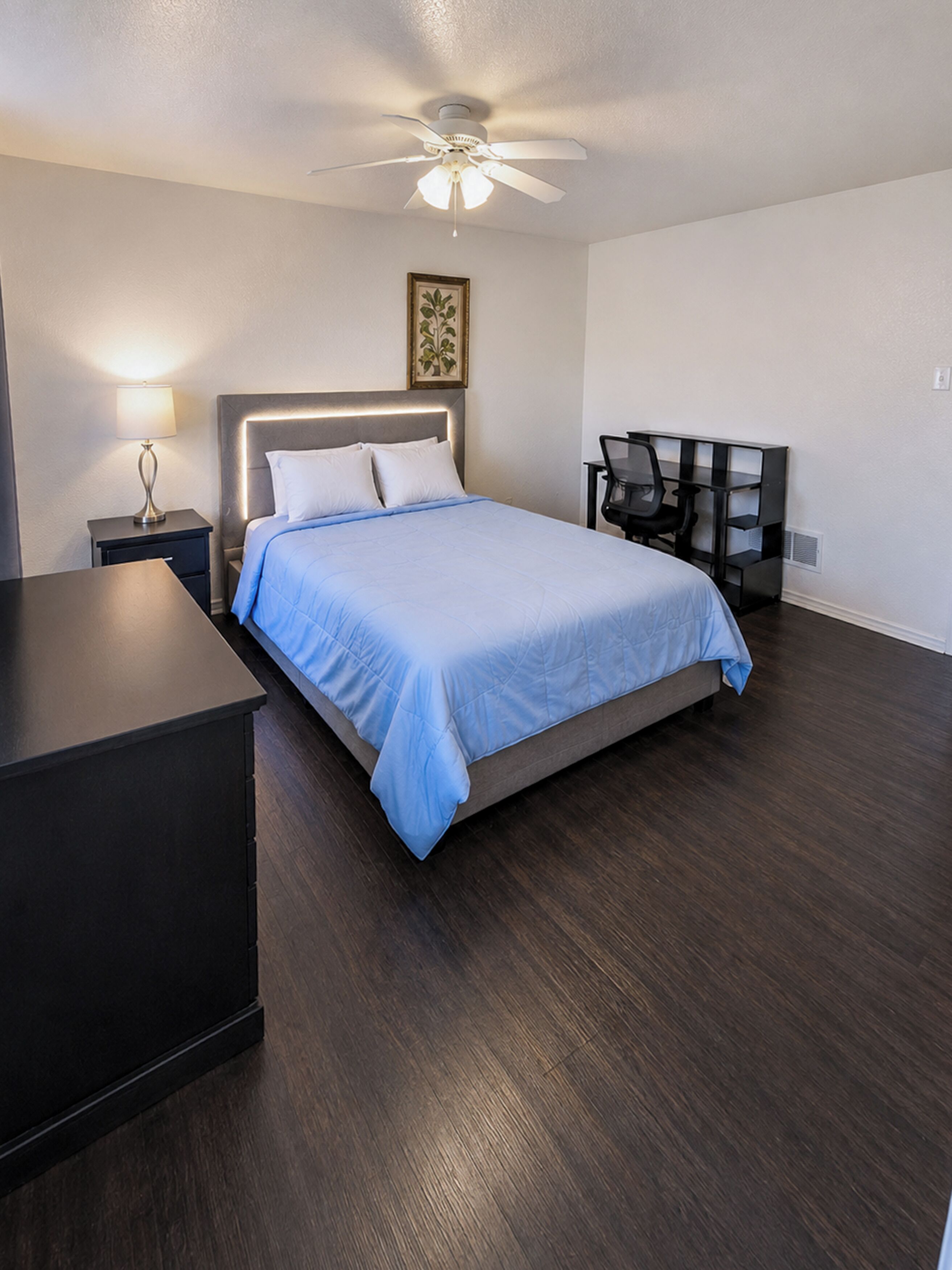Une chambre à coucher comprenant un lit, un bureau, une chaise, une table de chevet, une lampe et un ventilateur de plafond.