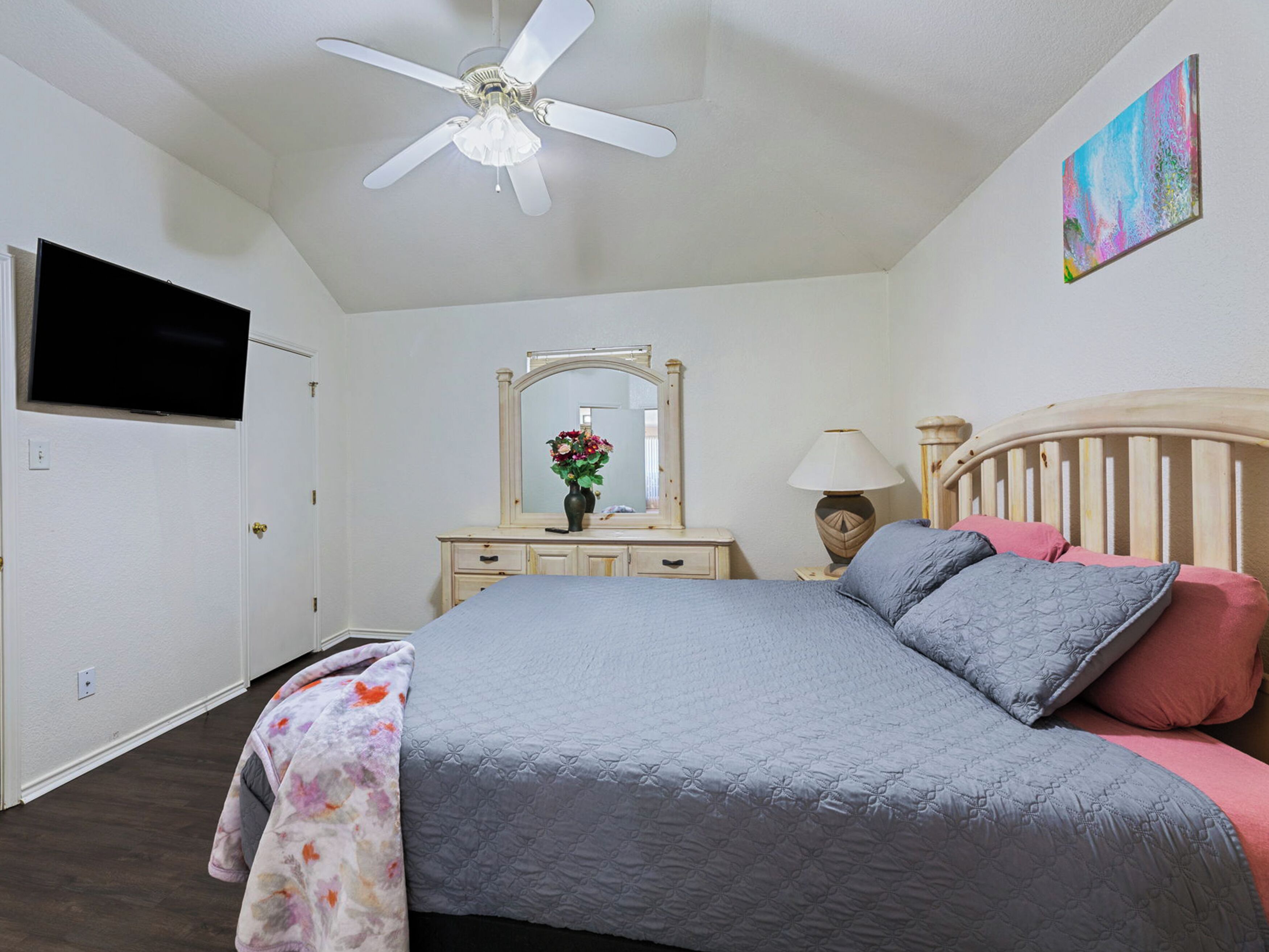 A bedroom with a bed, a ceiling fan, a dresser, a mirror, a TV, and a painting on the wall.