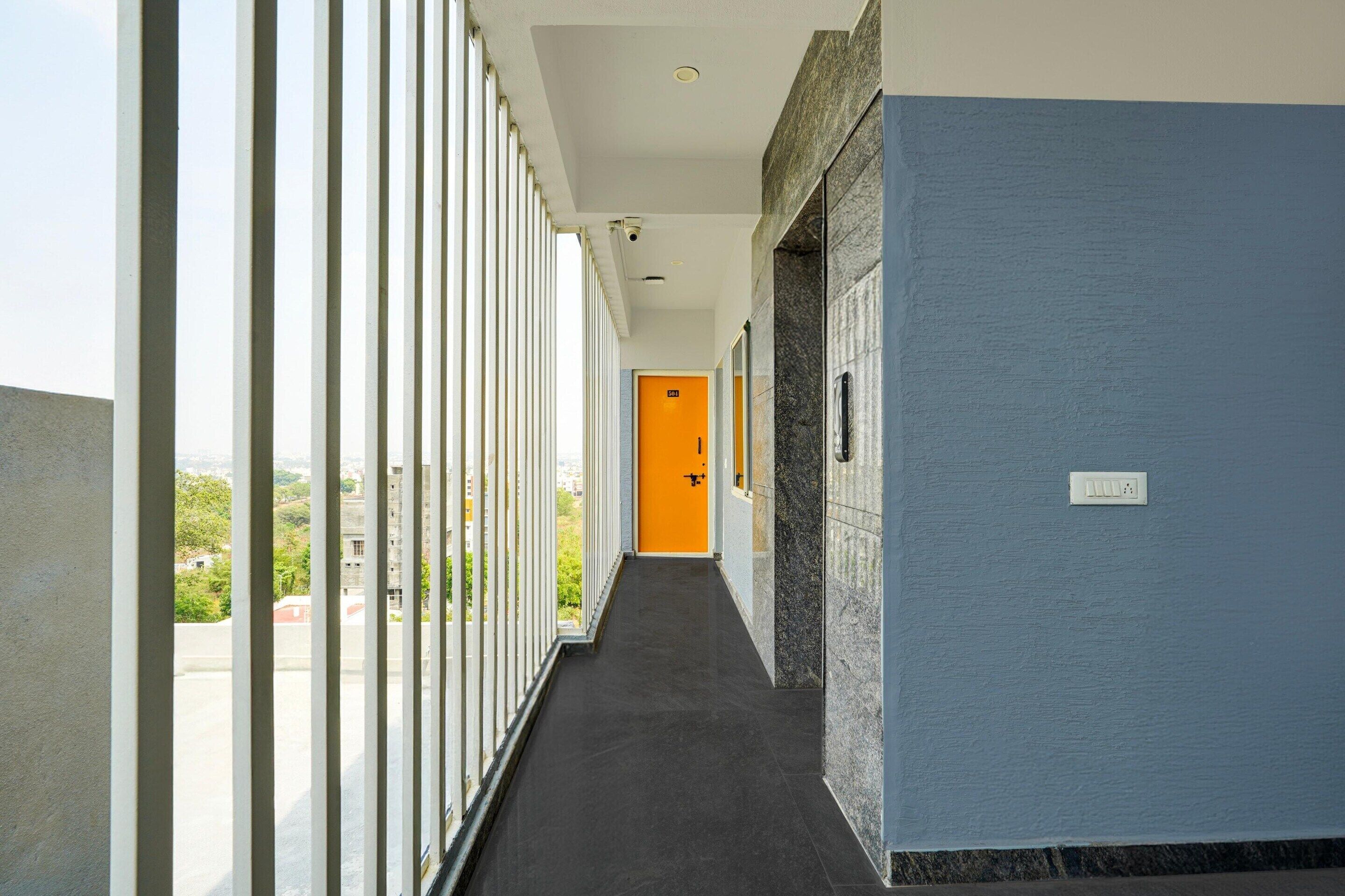 A corridor with a view of buildings outside, a door with a number, and a security camera on the ceiling.