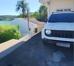 A white SUV parked on a narrow driveway with a view of a body of water and greenery. - Vista incrível do mar e acesso exclusivo à praia (Governador Celso Ramos)