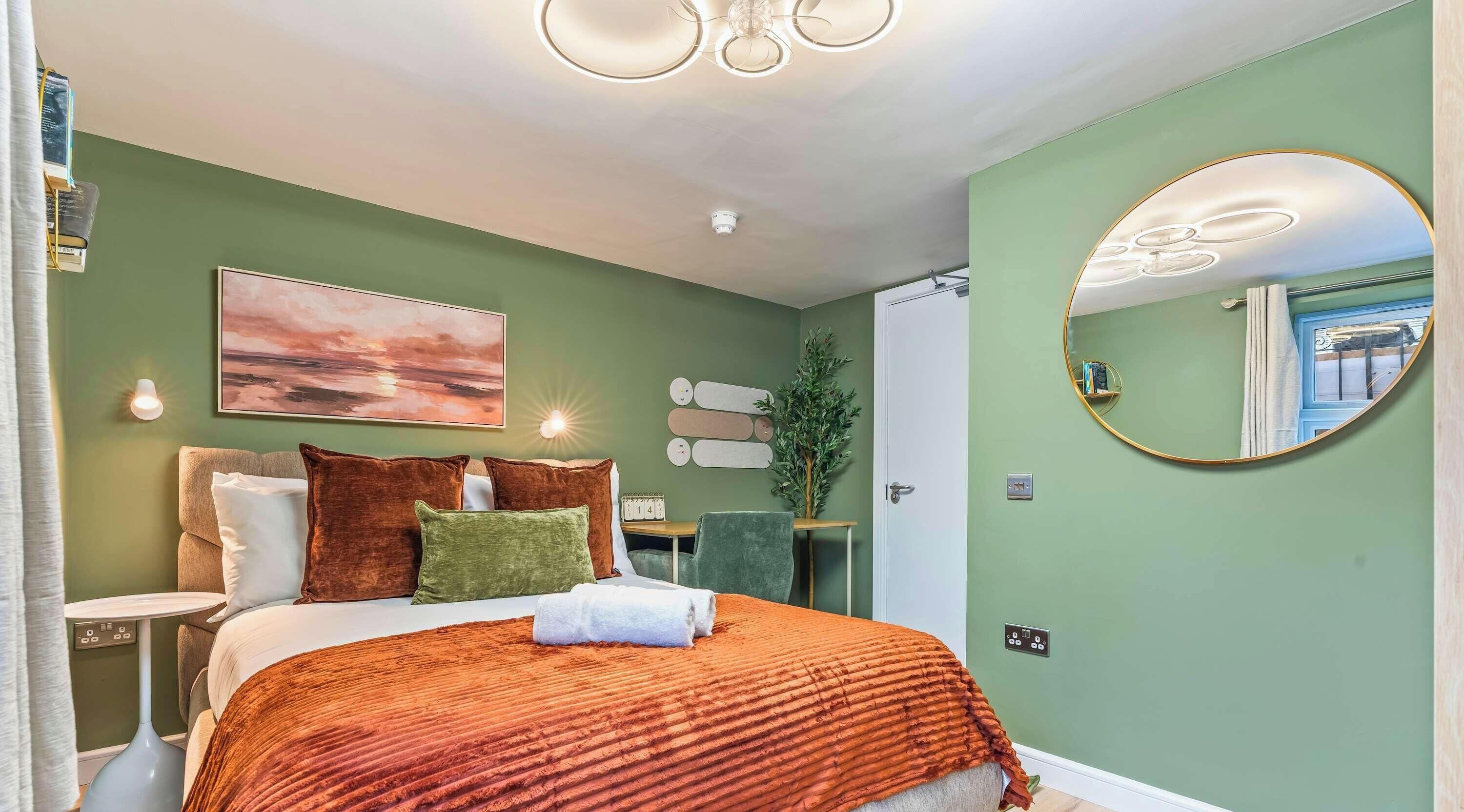 A well-lit bedroom with a bed, pillows, a bedside table, a mirror, and a wall-mounted light fixture.