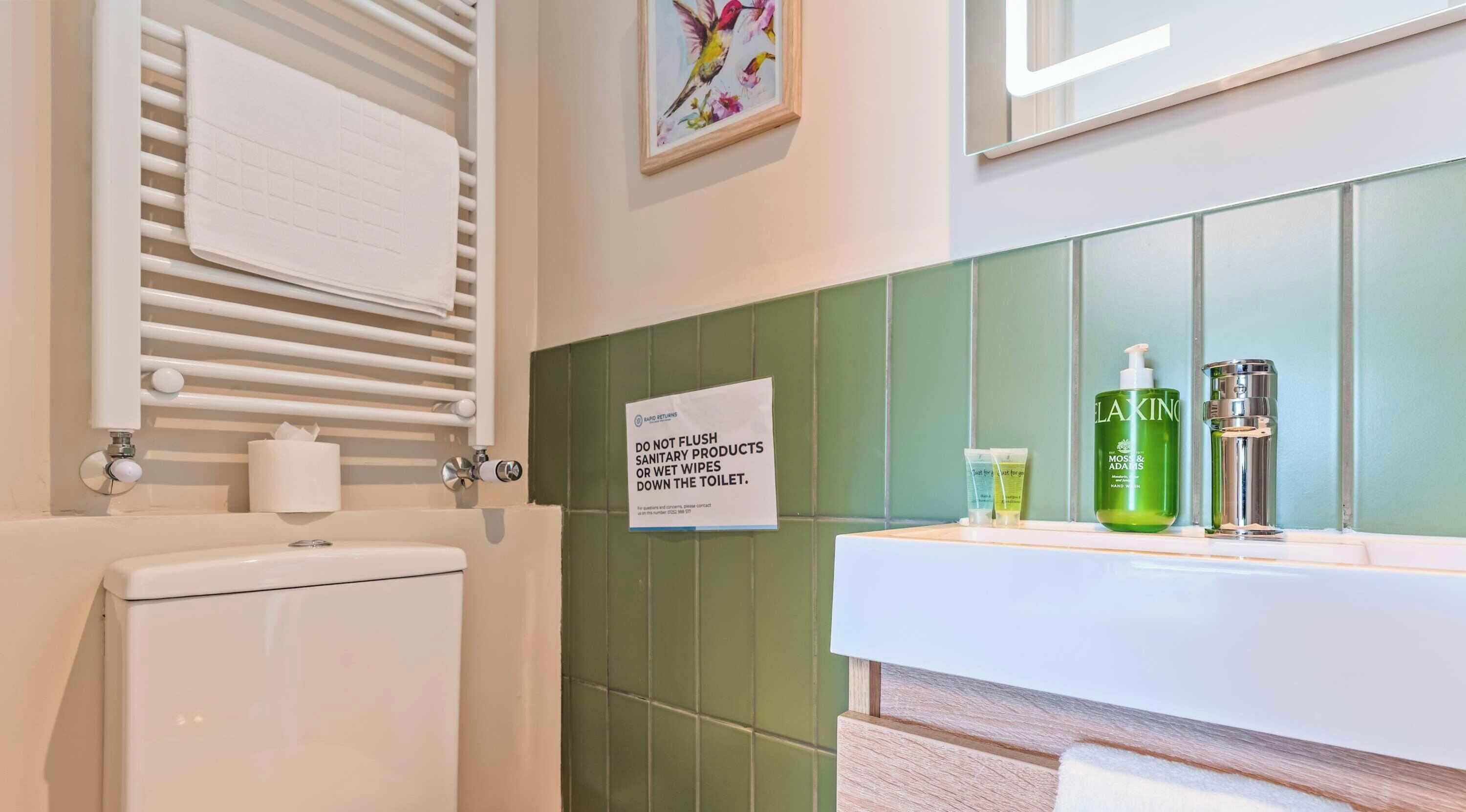 A bathroom with a toilet, a towel rack, a green tiled wall, a sink with toiletries, and a sign instructing not to flush sanitary products down the toilet.