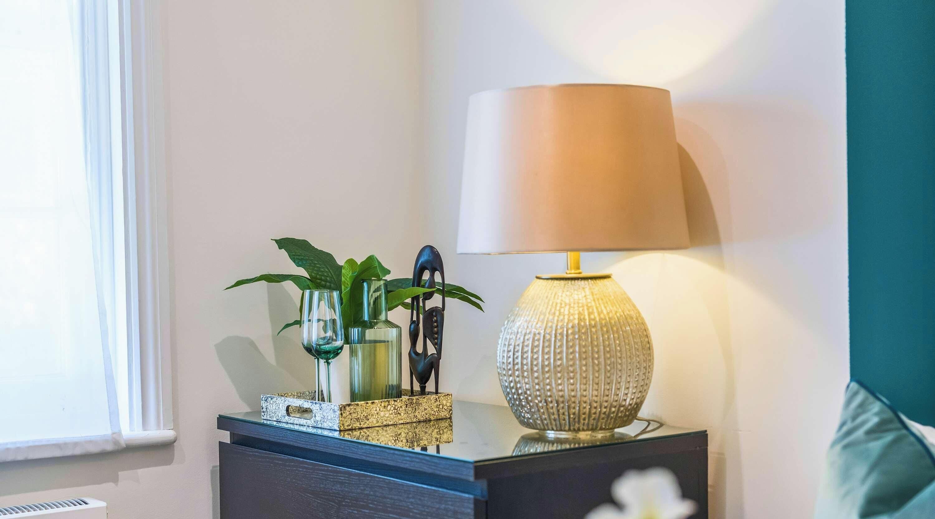 A bedside table with a lamp, decorative glass, and a plant.