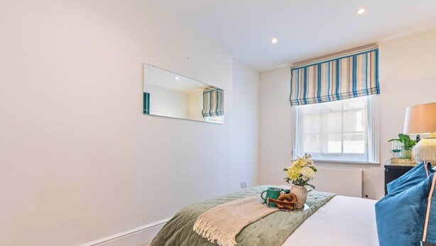 Une chambre à coucher avec un lit, une table de chevet avec une lampe, une fenêtre avec des rideaux et un miroir au mur.