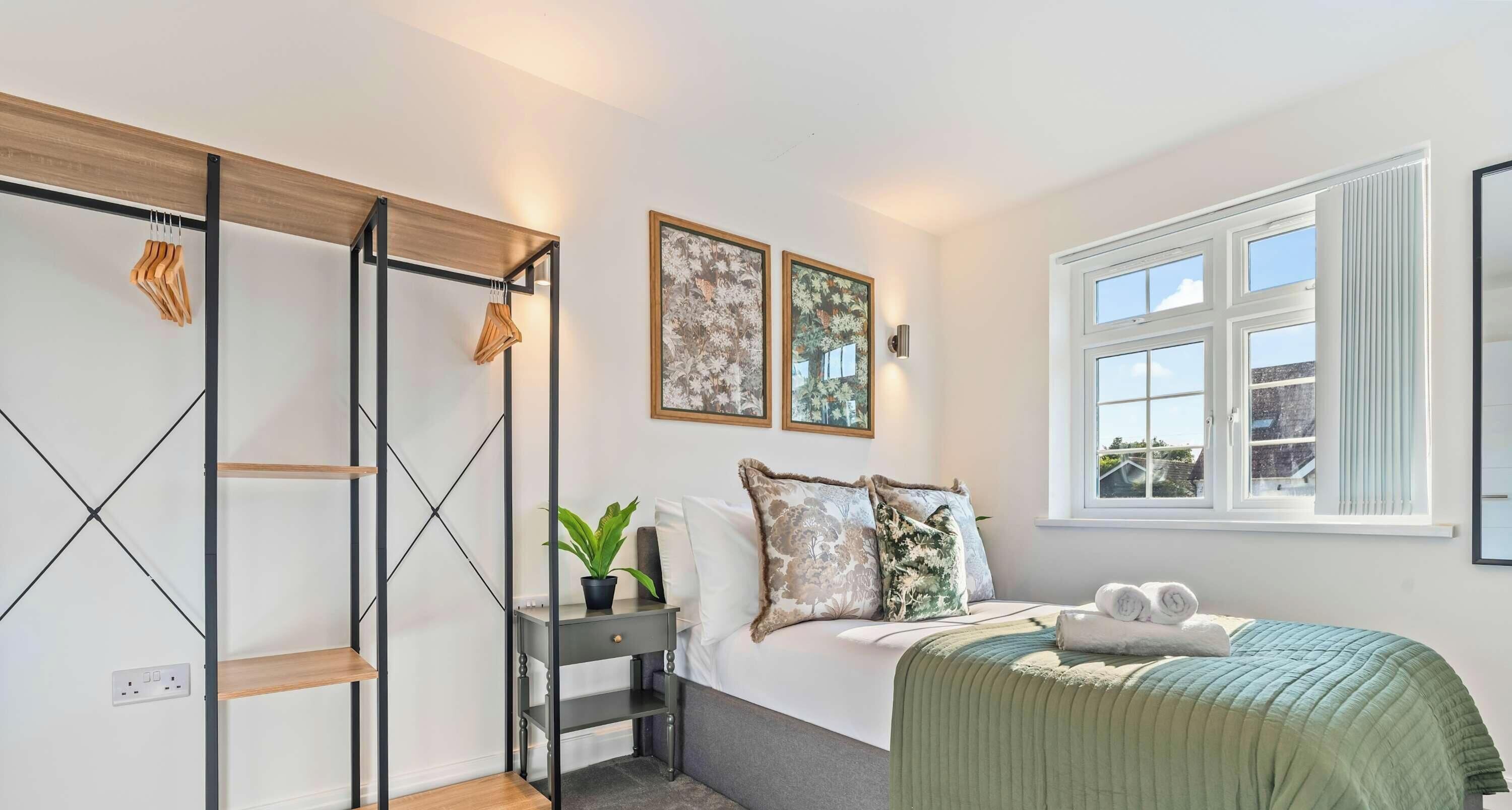 A bedroom with a bed, a bedside table with a plant, a wall-mounted shelf, and a window with blinds.