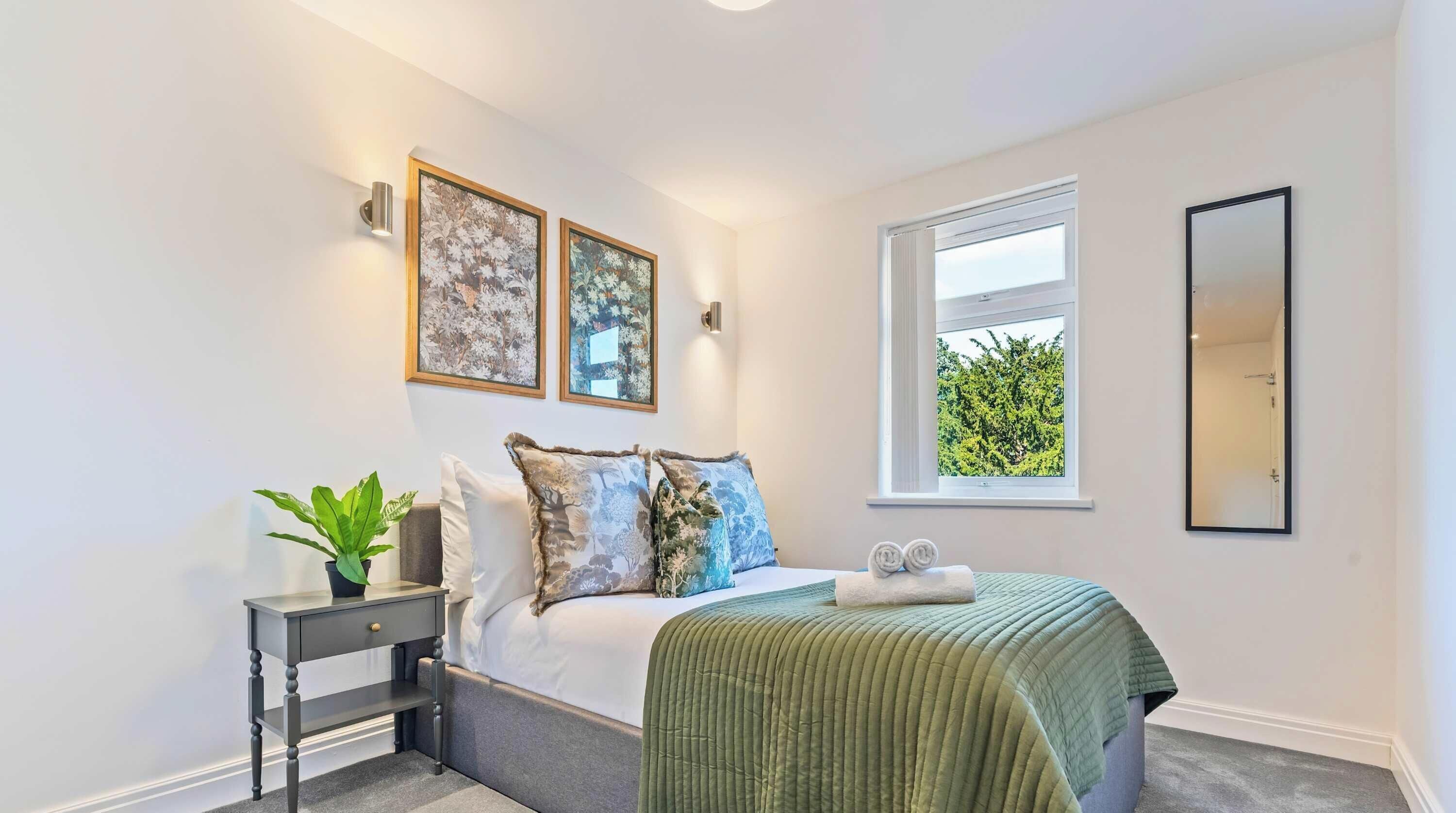 Un dormitorio con cama, una mesita de noche con una planta, dos lámparas de pared y una ventana con vistas a árboles.