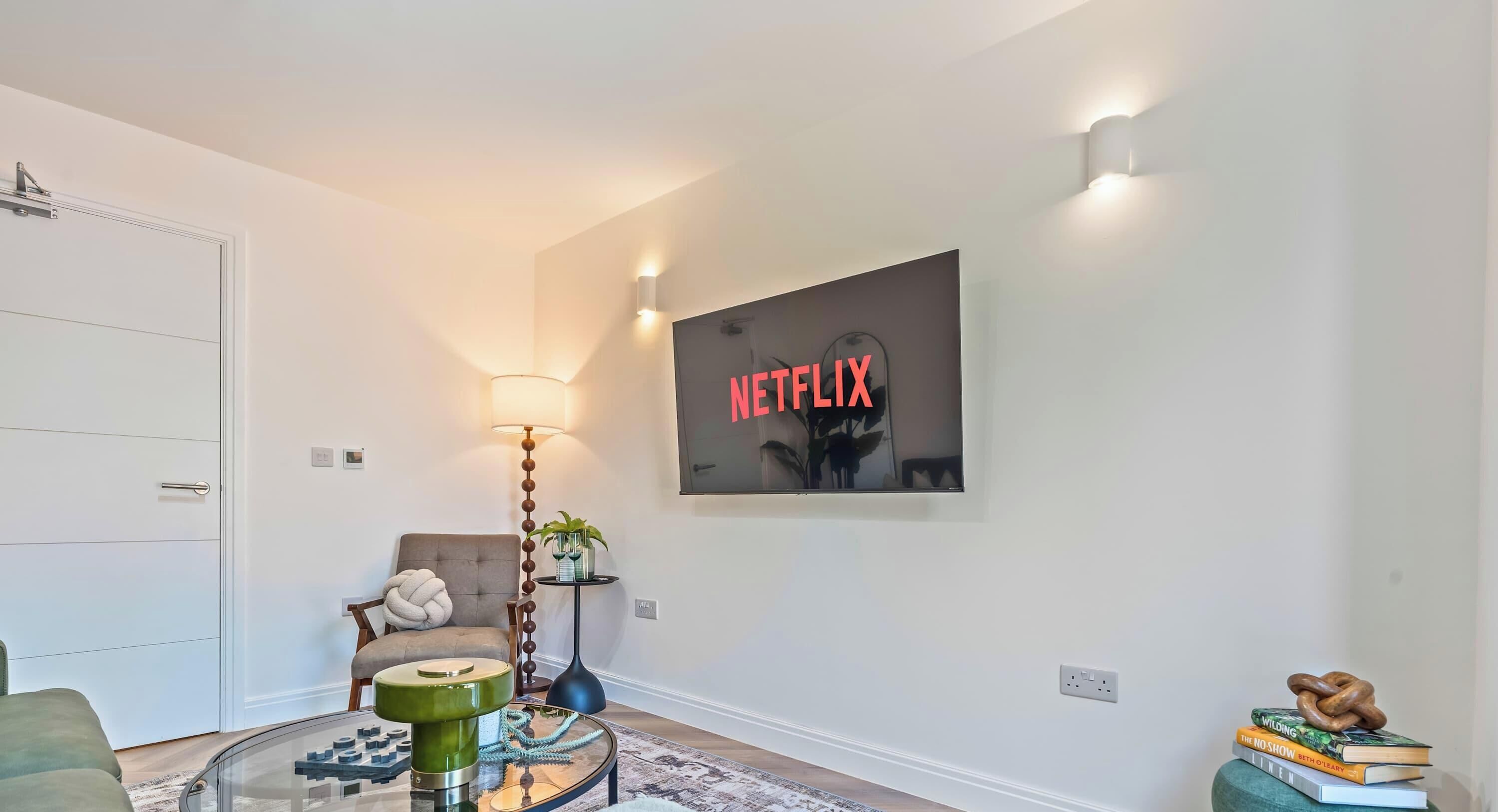 Una sala de estar moderna con un sofá, una mesa de centro, una lámpara, un televisor montado en la pared mostrando 'NETFLIX' y una pequeña estantería con libros.
