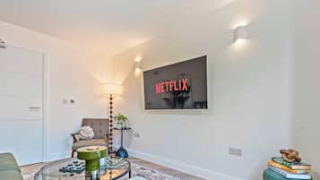 Una sala de estar moderna con un sofá, una mesa de centro, una lámpara, un televisor montado en la pared mostrando 'NETFLIX' y una pequeña estantería con libros.