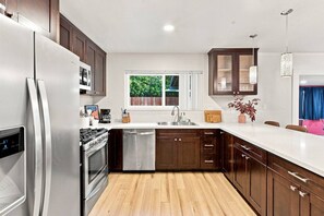 A modern kitchen with stainless steel appliances, wooden cabinets, a large window, and a view of greenery outside. - SDSU Splash Royale 6BR - 12 Bed - Pool & Spa - Game Room - AC - Washer & Dryer (San Diego)