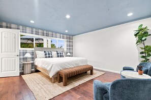 A bedroom with a bed, bedside tables, a bench, a chair, a potted plant, and a window with a view. - SDSU Splash Royale 6BR - 12 Bed - Pool & Spa - Game Room - AC - Washer & Dryer (San Diego)