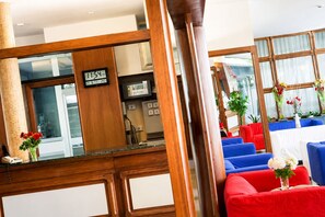 Un vestíbulo de hotel con mostrador de recepción de madera, un reloj digital y zonas de estar en rojo y azul.