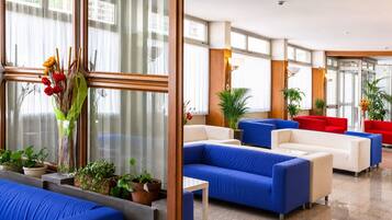 Un hall de hotel con sofás azules y rojos, plantas en macetas y grandes ventanales.