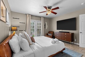 Una habitación de hotel con una cama, ventilador de techo, televisor, cómoda y ventana con persianas.