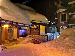 A snow-covered street with buildings, a stone wall, and a wooden structure with a sign. - Very comfortable apartment in Belle-Plagne, 50 m² (La Plagne Tarentaise)