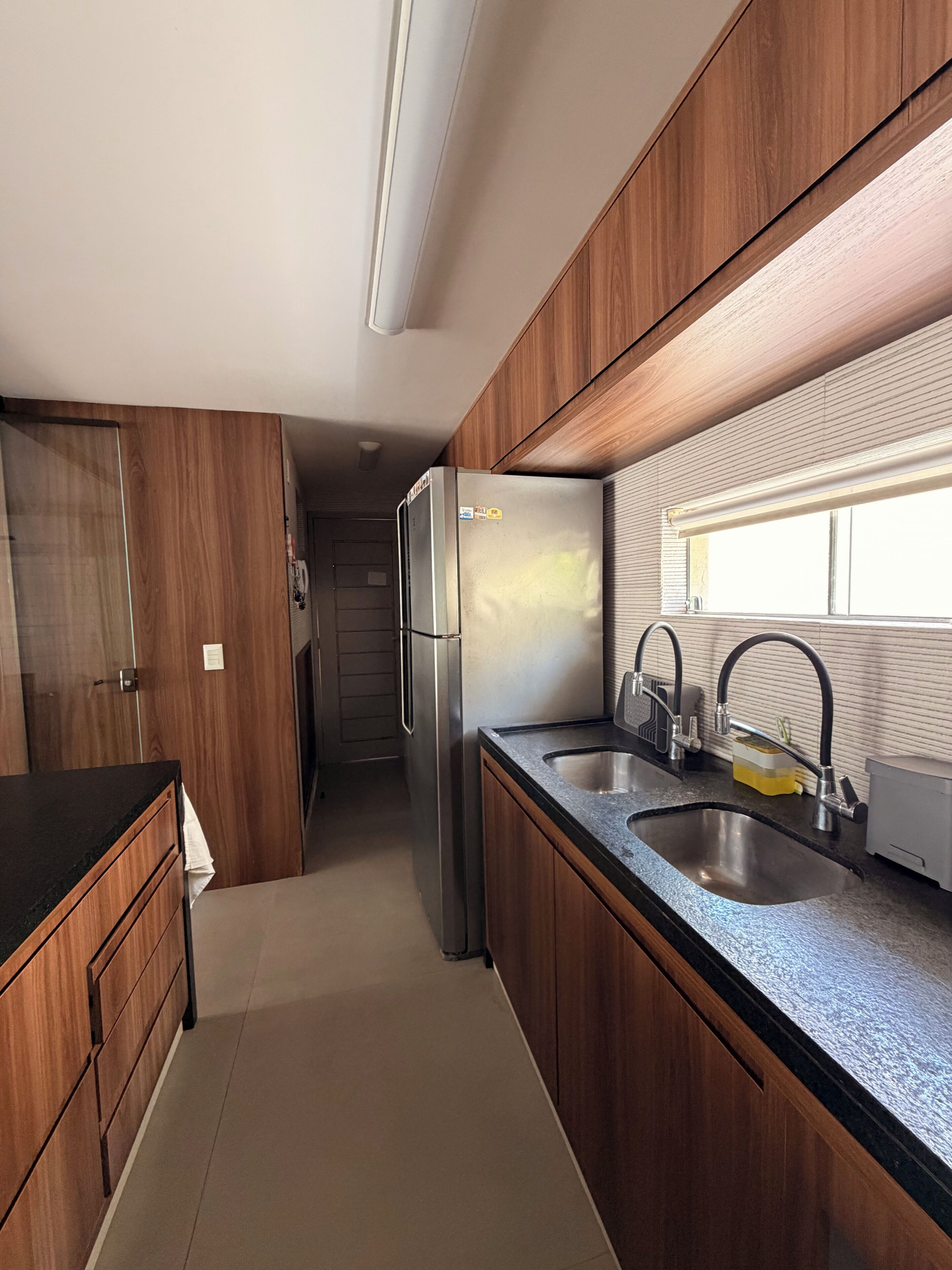 A compact kitchen with wooden cabinets, a stainless steel refrigerator, a double sink, and a window providing natural light.