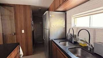 A compact kitchen with wooden cabinets, a stainless steel refrigerator, a double sink, and a window providing natural light.