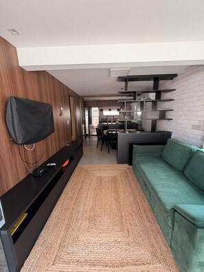 Un soggiorno moderno con TV a schermo piatto montata a parete, un divano verde, una parete d'accento in legno e un angolo cottura sullo sfondo.