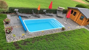 A rectangular swimming pool with a stone border, surrounded by a patio with lounge chairs and umbrellas.