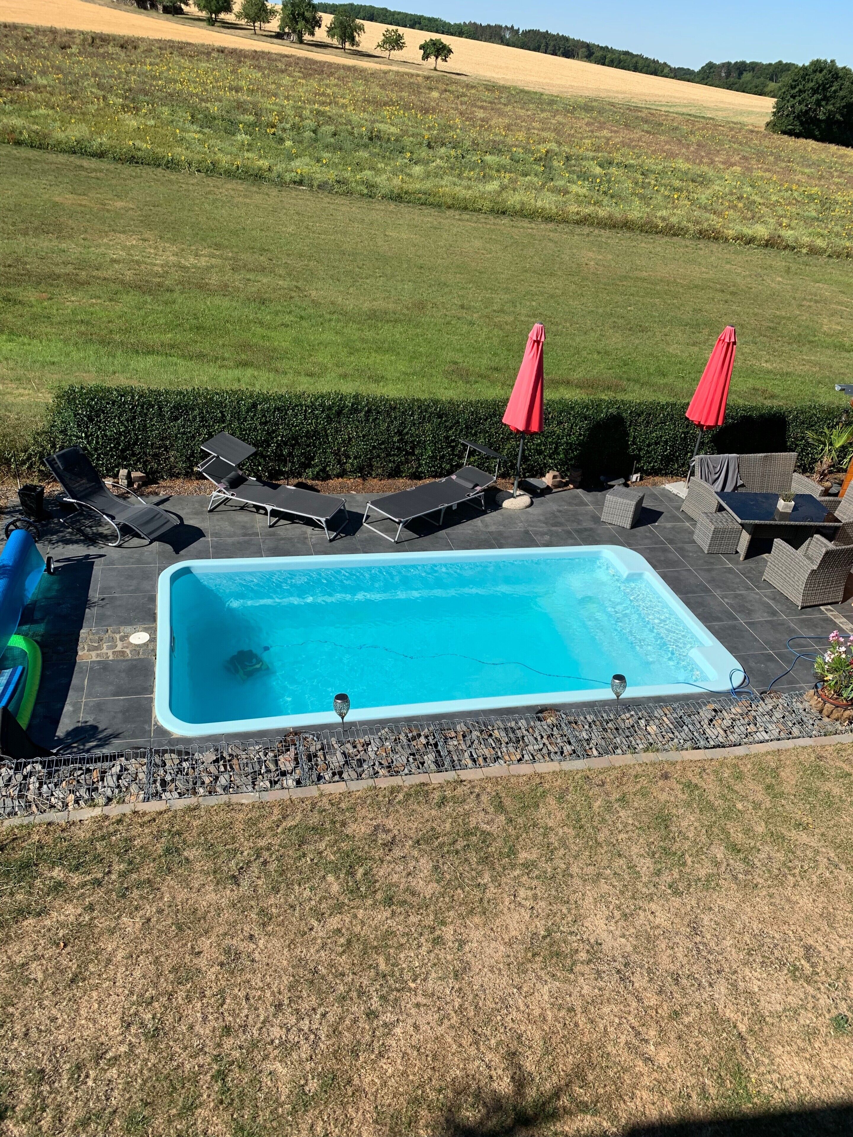 Ein rechteckiger Swimmingpool mit einer gefliesten Liegefläche, umgeben von Liegestühlen und einem roten Sonnenschirm.