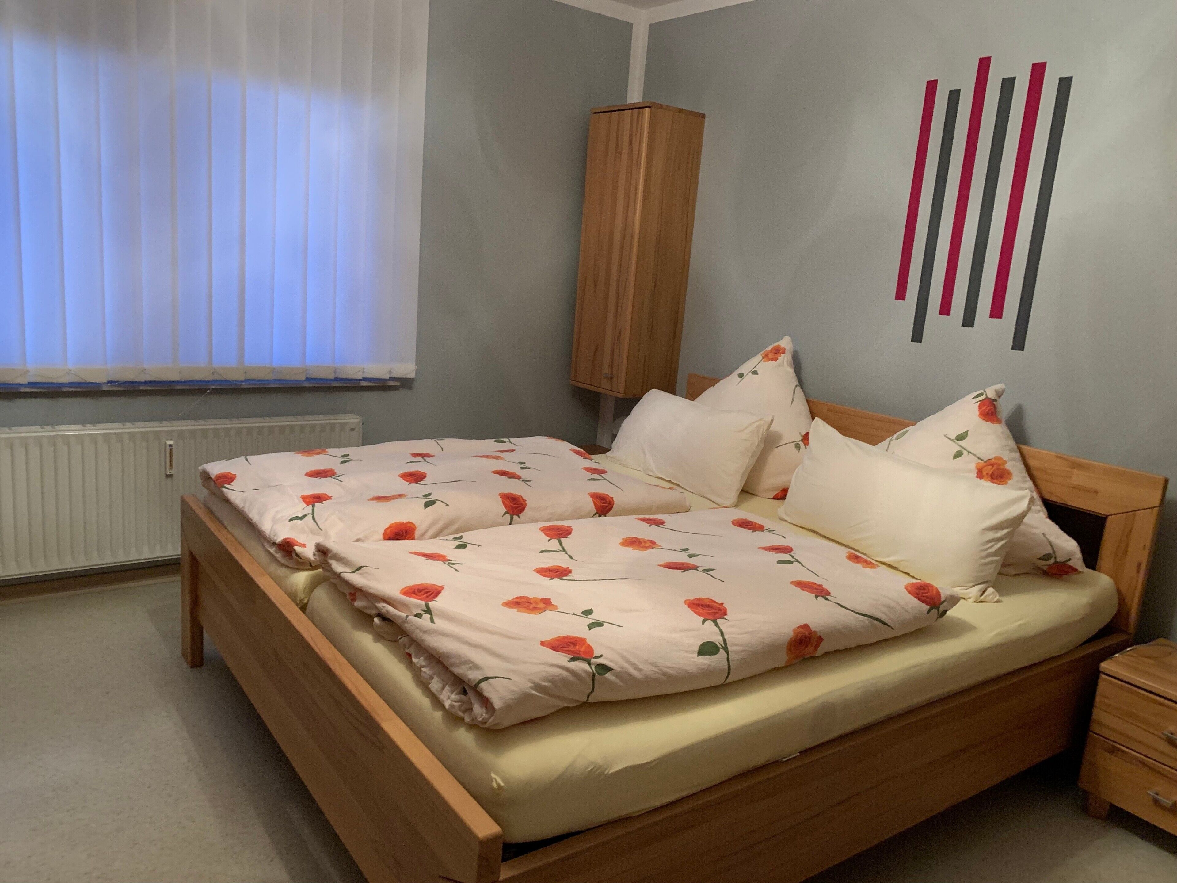 Una camera da letto con un letto di legno, biancheria da letto bianca con motivo floreale, un comodino in legno, un termosifone e una finestra con persiane.