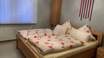 Una camera da letto con un letto di legno, biancheria da letto bianca con motivo floreale, un comodino in legno, un termosifone e una finestra con persiane.