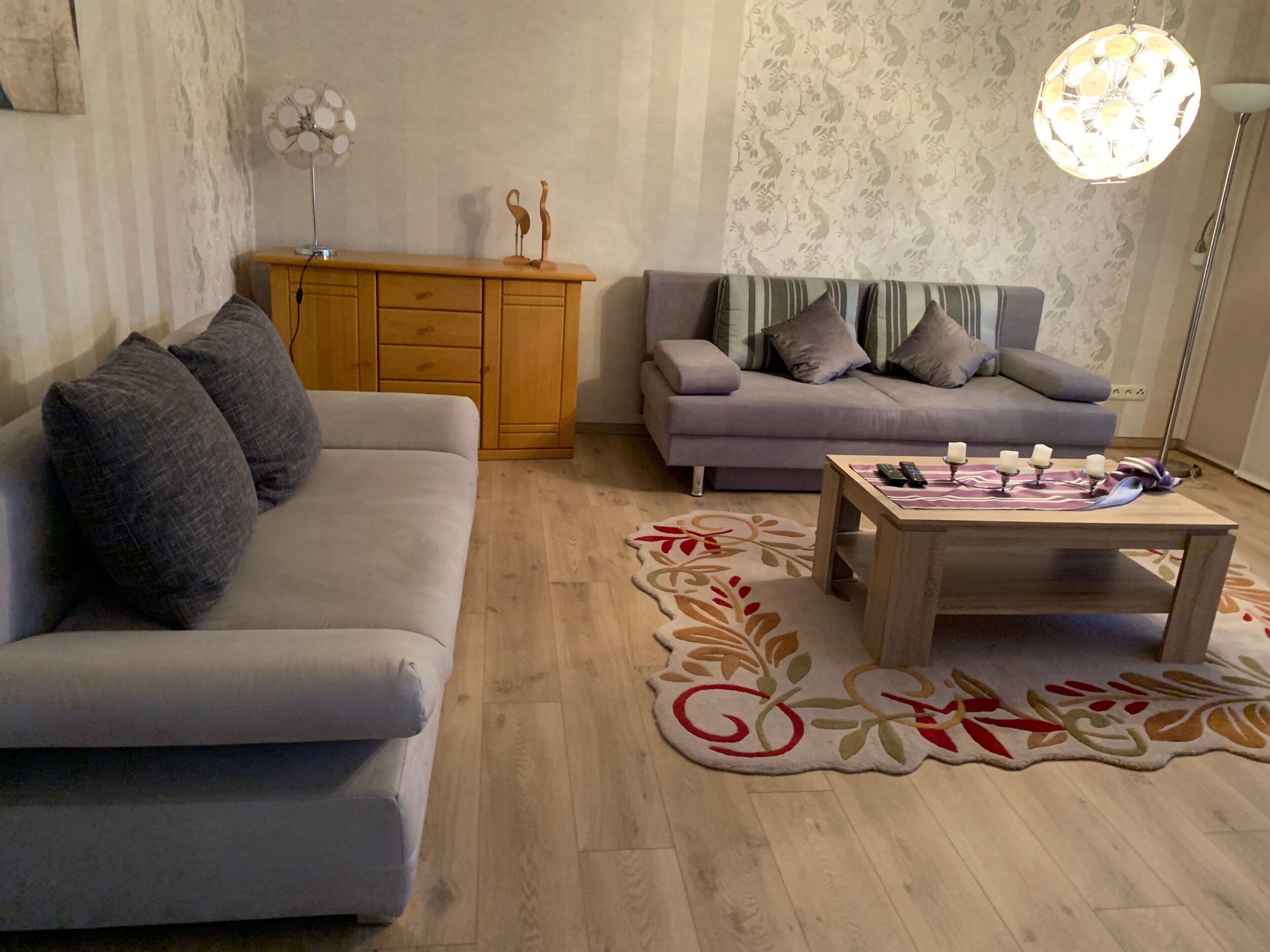 Ein Wohnzimmer mit einer Couch, einem Couchtisch mit Fernbedienungen, einem gemusterten Teppich und einer Wanddekoration.