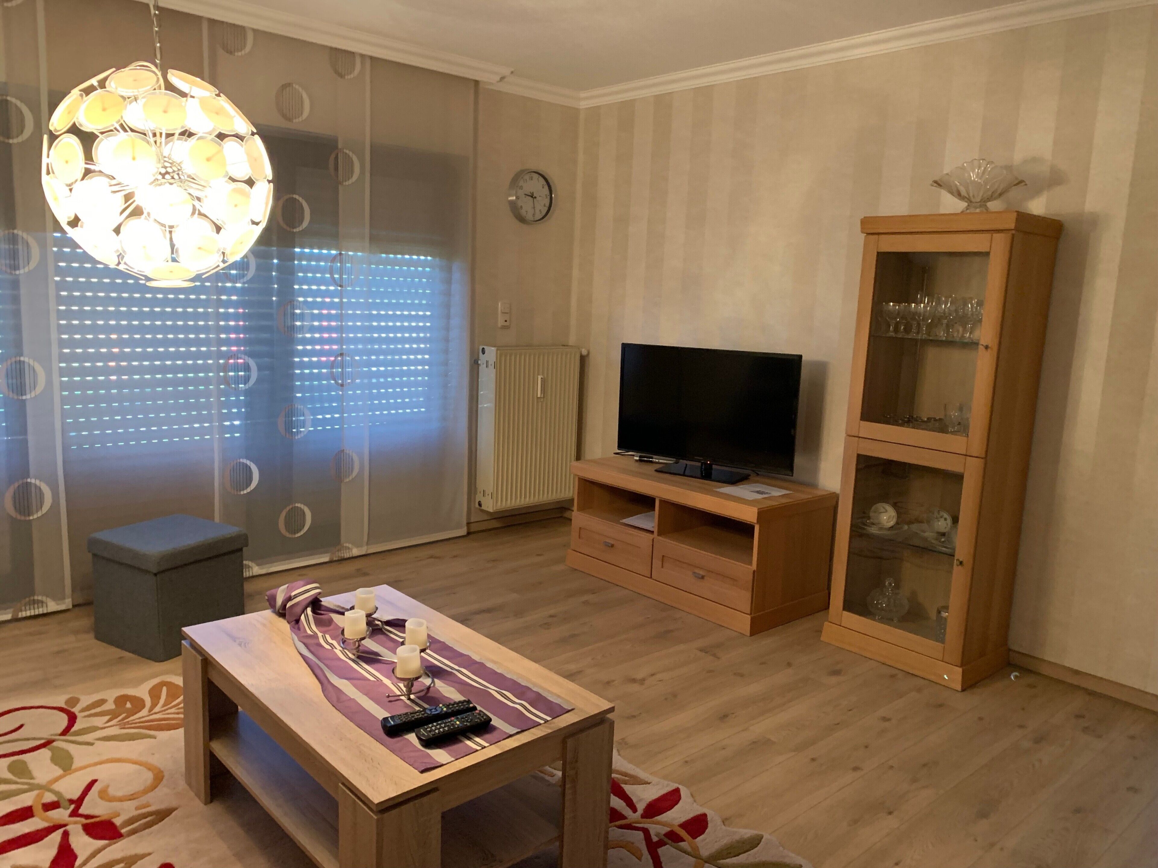 A living room with a wooden TV stand, a glass cabinet, a table with remote controls, and a decorative light fixture.
