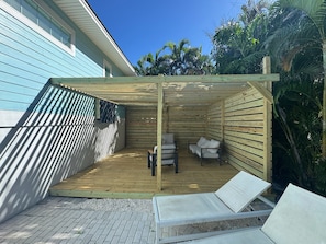 Ett takförsett utomhusområde med sittplatser, bestående av två solstolar och ett bord, omgivet av en träpergola.
