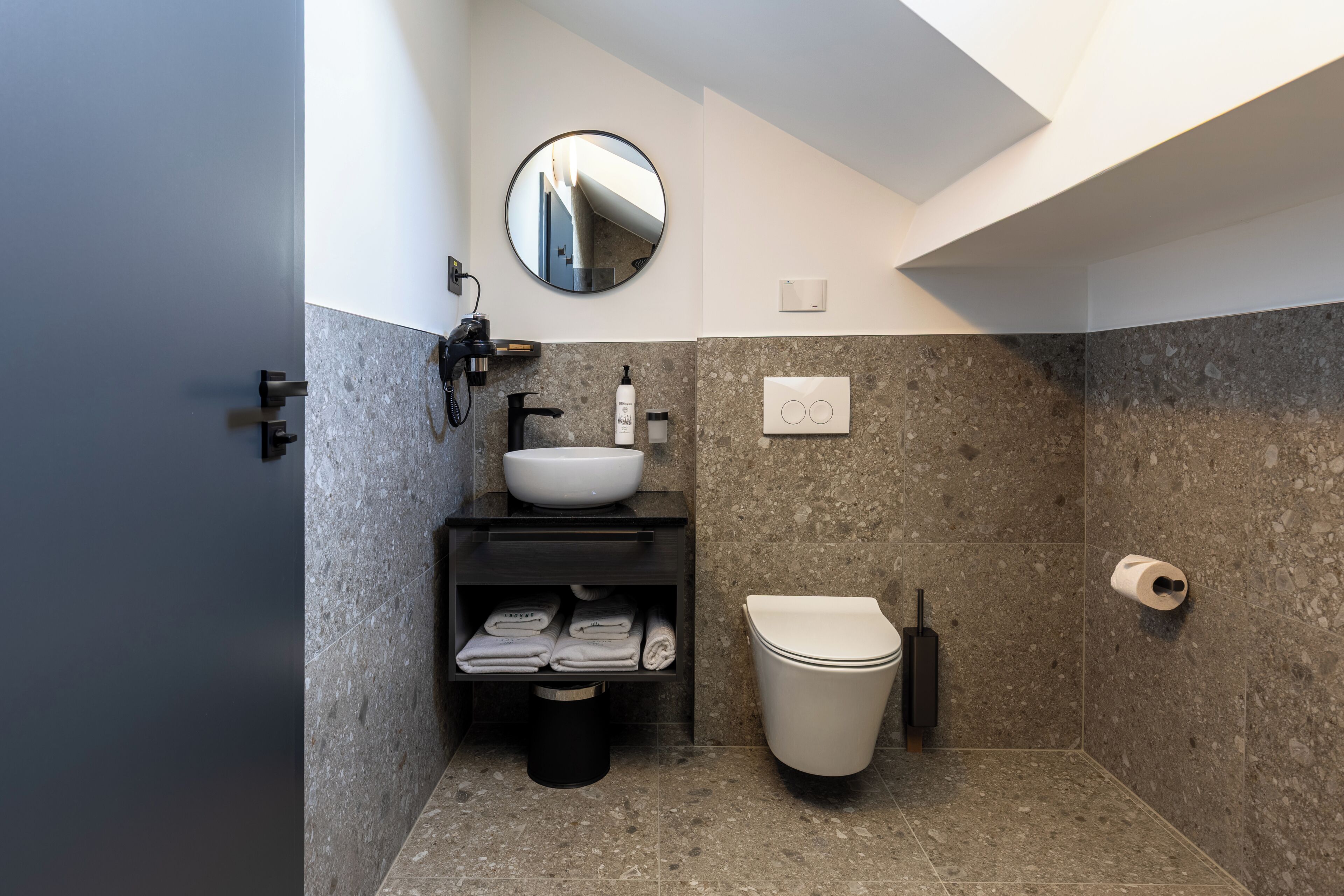 A modern bathroom with a built-in sink, a toilet, a shelf with folded towels, a trash bin, and a toilet paper holder.