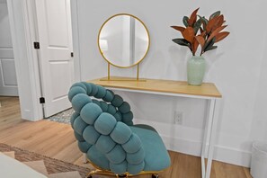 Un fauteuil recouvert d’un tissu de couleur turquoise avec un design original et torsadé, un miroir rond posé sur un pied, et un vase contenant des feuilles vertes, placé sur une étagère en bois.