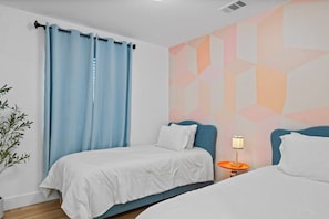 A hotel room with a bed, a nightstand with a lamp, a window with blue curtains, and a geometric wall design. - Pixel Palace•Spa•Gameroom•Theatre•Sleeps 14 (San Diego)