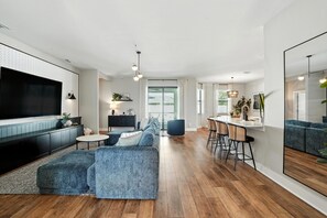 Ein modernes Wohnzimmer mit einem großen Flachbildfernseher, einer Sofagarnitur, einem Essbereich mit Barhocker und einer durch eine offene Tür einsehbarer Küche.