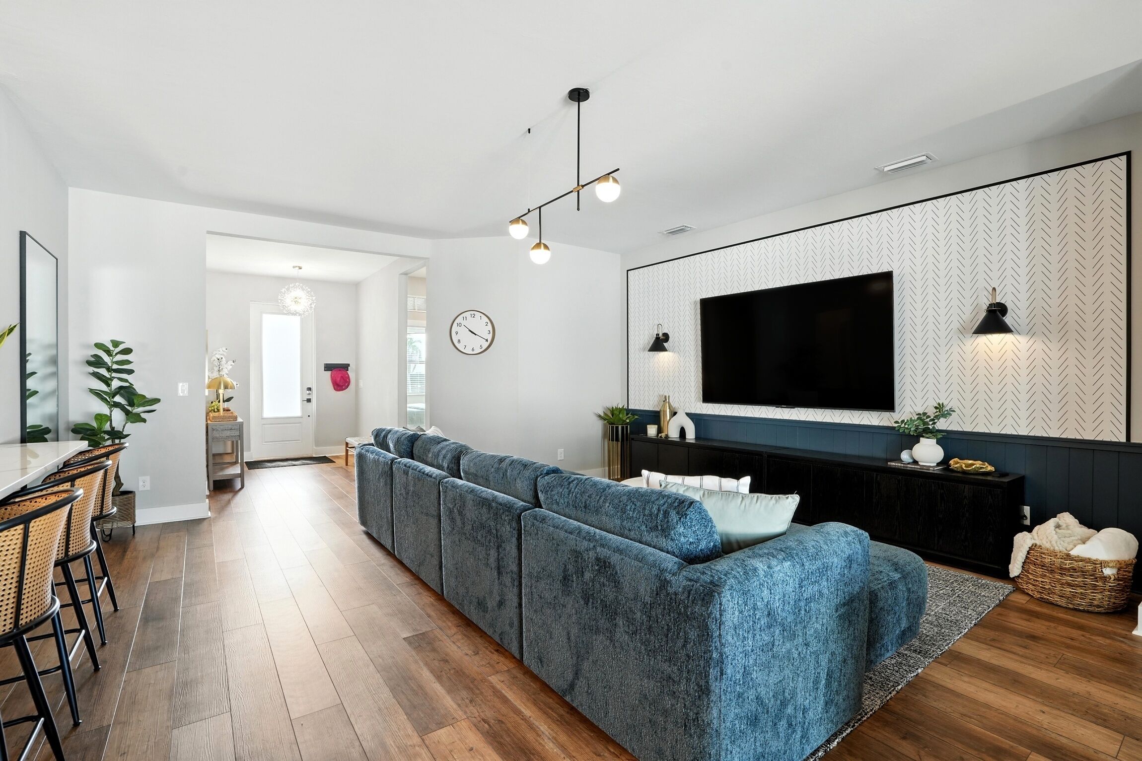 A modern living room with a sectional sofa, a flat-screen TV mounted on a patterned wall, and a dining area with a table and chairs.