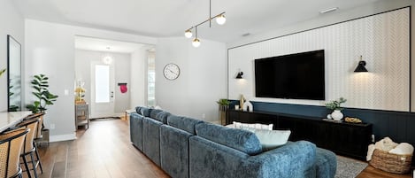 Un salon moderne avec un canapé d’angle, un téléviseur à écran plat fixé à un mur orné de motifs, et un coin repas comprenant une table et des chaises.