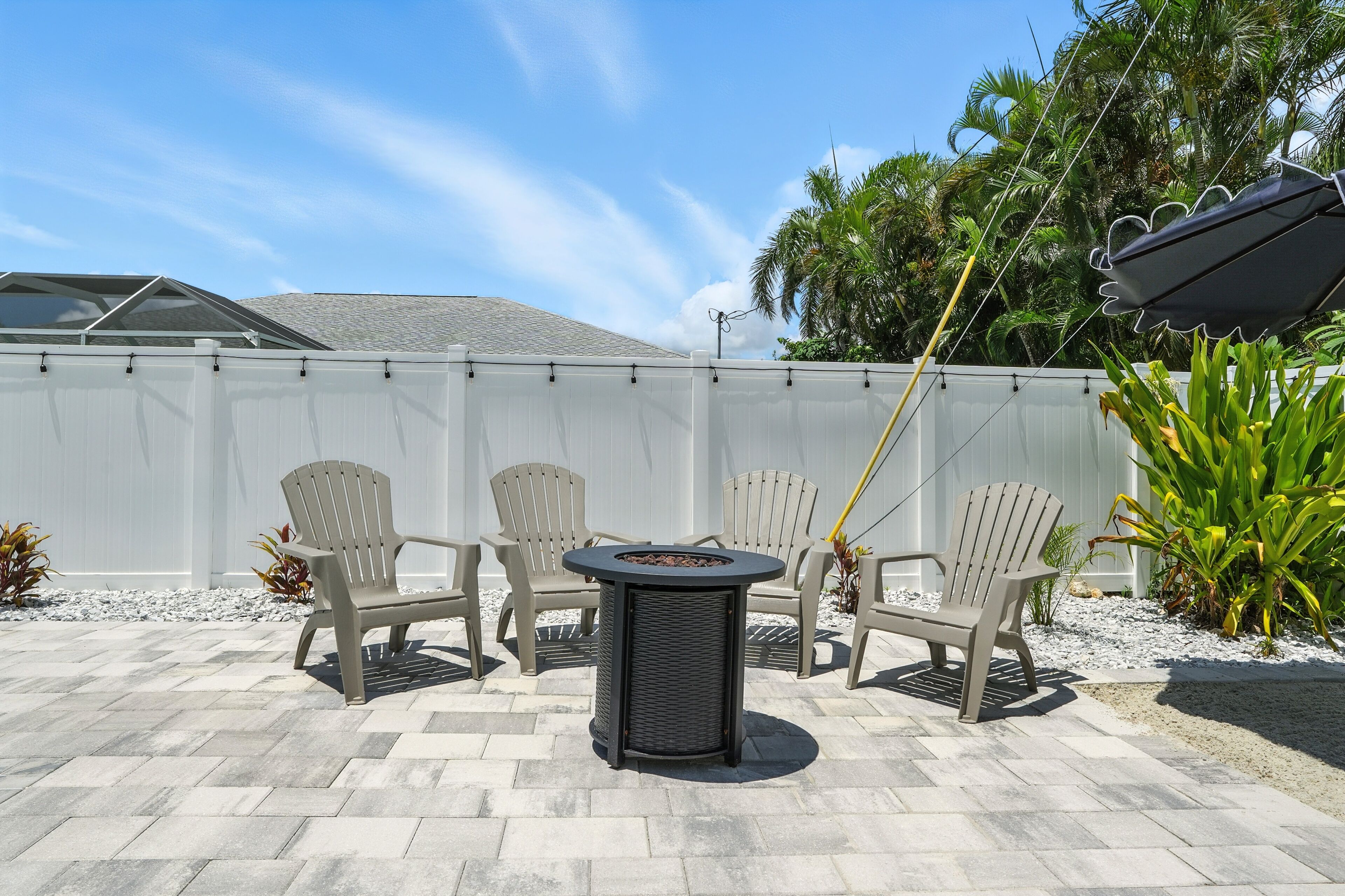 A patio area with a table and four chairs, a white fence, and tropical plants.