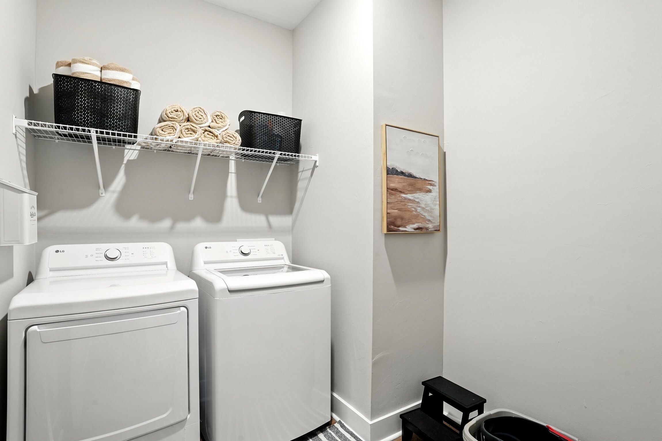 A laundry area with two white washing machines, a shelf with towels, and a framed picture on the wall.