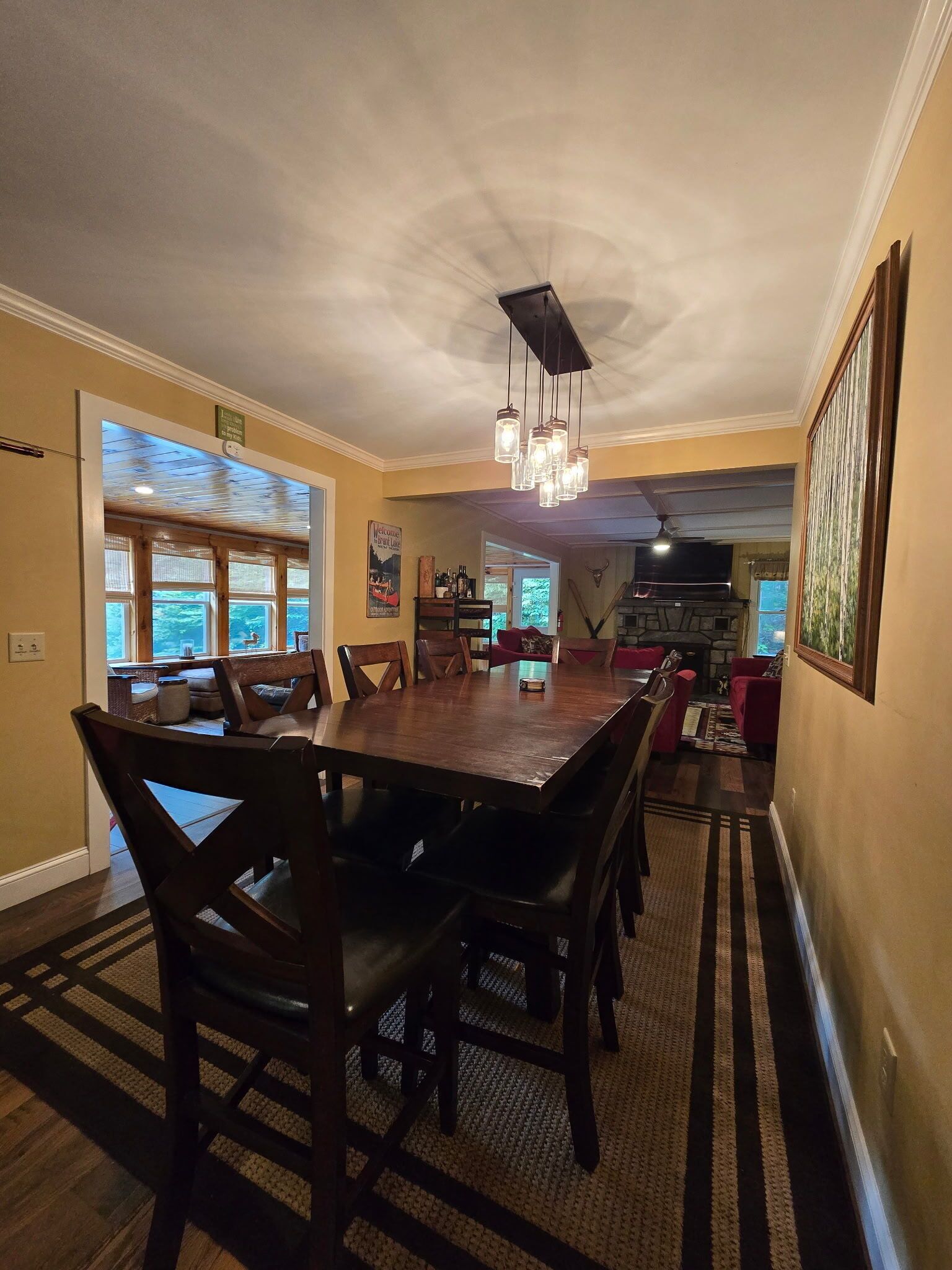 A dining area with a large wooden table, multiple chairs, a hanging light fixture, and a fireplace in the background.